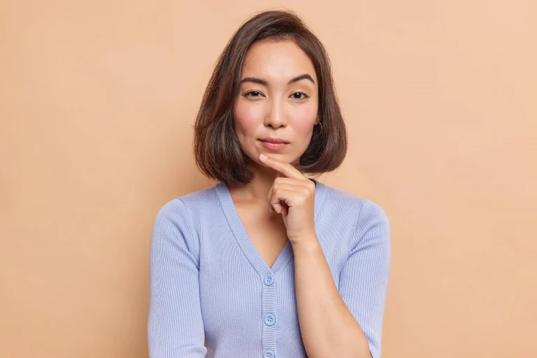 portrait-serious-dark-haired-asian-woman-keeps-finger-chin-looks-mysteriously-front-considers-someth